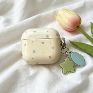 May include: A cream-colored protective case for wireless earbuds featuring a pattern of small, multicolored stars. A metal ring is attached to the side, holding a light green star-shaped charm and a small, oval, light blue acrylic tag.