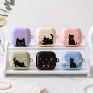 May include: Six square protective cases for wireless earbuds, each featuring a black cat illustration. The cases are colored lavender, cream, pink, sage green, black, and light blue, and are displayed on a two-tiered white shelf.