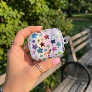 Pode incluir: Uma capa protetora para auriculares sem fios com acabamento hologr&aacute;fico iridescente, decorada com um padr&atilde;o de estrelas brilhantes multicoloridas em tons de azul, roxo, rosa e dourado, incluindo uma argola lateral para fixa&ccedil;&atilde;o.