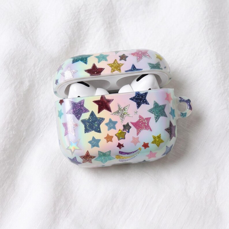 Pode incluir: Uma capa protetora para auriculares sem fios com um fundo de gradiente arco-&iacute;ris pastel coberto por um padr&atilde;o de estrelas brilhantes multicoloridas. A capa est&aacute; aberta, revelando auriculares brancos no interior, e inclui uma pequena argola lateral.