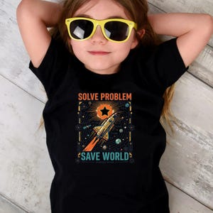 May include: A black t-shirt featuring a graphic print of a rocket ship launching into space, surrounded by scientific symbols, planets, and a star. The text reads SOLVE PROBLEM, SAVE WORLD, and SCIENCE SAVES in orange and teal lettering.