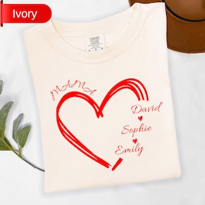 May include: Ivory-colored t-shirt with a red heart design. The heart contains the names "Mama", "David", "Sophie", and "Emily". The shirt is made of a soft material and is labeled "Comfort Colors".