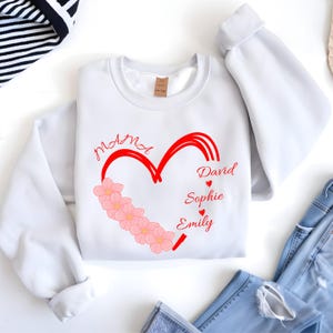 May include: White sweatshirt with a red heart design. The heart is filled with pink flowers and the names "David", "Sophie", and "Emily" are written in red. The word "Mama" is also written in red.