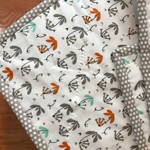 K&ouml;nnte beinhalten: Eine wei&szlig;e Steppdecke mit einem Blumenmuster in Braun-, Orange- und Blaut&ouml;nen. Die Steppdecke hat einen grauen und wei&szlig;en Tupfenrand.