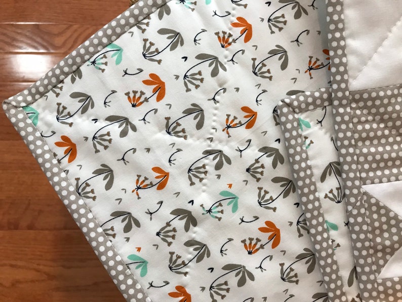 K&ouml;nnte beinhalten: Eine wei&szlig;e Steppdecke mit einem Blumenmuster in Braun-, Orange- und Blaut&ouml;nen. Die Steppdecke hat einen grauen und wei&szlig;en Tupfenrand.