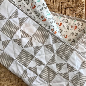 K&ouml;nnte beinhalten: Eine wei&szlig;e und graue Steppdecke mit einem geometrischen Muster aus Dreiecken und Punkten. Die Steppdecke ist in der H&auml;lfte gefaltet und liegt auf einer geflochtenen Weidenoberfl&auml;che.