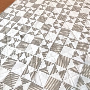 K&ouml;nnte beinhalten: Eine wei&szlig;e und graue Steppdecke mit einem geometrischen Muster aus Quadraten und Dreiecken. Die Steppdecke besteht aus zwei Stoffen, einem mit wei&szlig;em Hintergrund und dem anderen mit grauem Hintergrund mit wei&szlig;en Punkten.