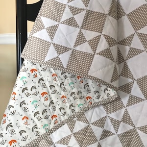 K&ouml;nnte beinhalten: Eine wei&szlig;e und braune Steppdecke mit einem geometrischen Muster aus wei&szlig;en Dreiecken auf braunem Grund mit wei&szlig;en Punkten. Die Steppdecke ist gefaltet und &uuml;ber einen Stuhl gelegt. Die R&uuml;ckseite der Steppdecke ist wei&szlig; mit einem Muster aus braunen und orangefarbenen Blumen.