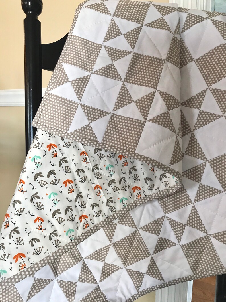 K&ouml;nnte beinhalten: Eine wei&szlig;e und braune Steppdecke mit einem geometrischen Muster aus wei&szlig;en Dreiecken auf braunem Grund mit wei&szlig;en Punkten. Die Steppdecke ist gefaltet und &uuml;ber einen Stuhl gelegt. Die R&uuml;ckseite der Steppdecke ist wei&szlig; mit einem Muster aus braunen und orangefarbenen Blumen.