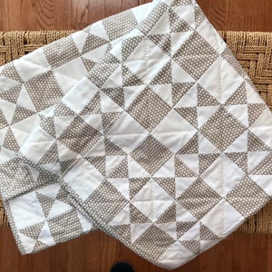 K&ouml;nnte beinhalten: Eine wei&szlig;e und graue Steppdecke mit einem geometrischen Muster aus Dreiecken und Quadraten. Die Steppdecke ist aus Baumwollstoff gefertigt und hat ein dezentes Punkte-Muster.