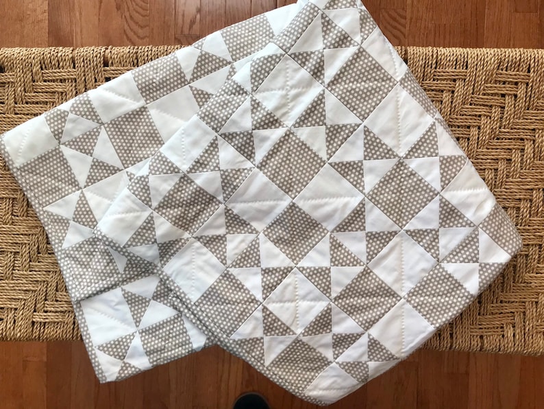 K&ouml;nnte beinhalten: Eine wei&szlig;e und graue Steppdecke mit einem geometrischen Muster aus Dreiecken und Quadraten. Die Steppdecke ist aus Baumwollstoff gefertigt und hat ein dezentes Punkte-Muster.