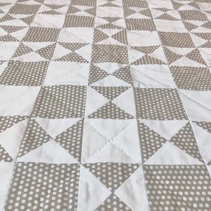K&ouml;nnte beinhalten: Eine Steppdecke aus wei&szlig;em und beigefarbenem Stoff mit einem Tupfenmuster. Die Steppdecke besteht aus Quadraten und Dreiecken, die ein geometrisches Muster bilden.