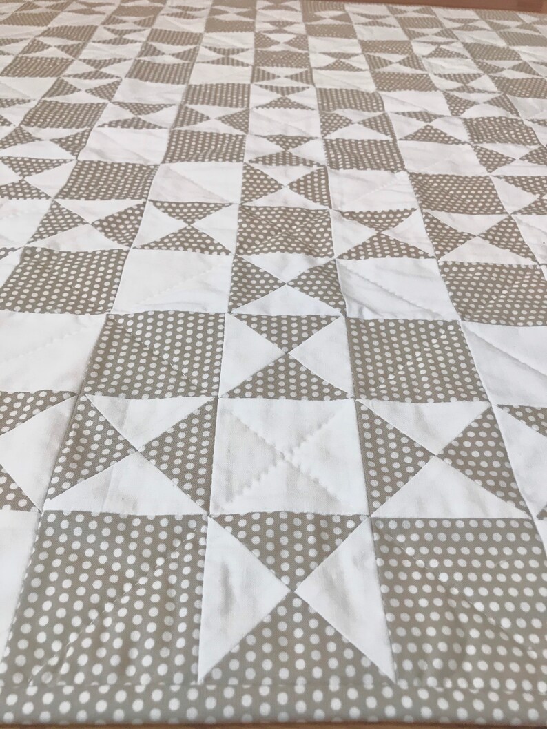 K&ouml;nnte beinhalten: Eine Steppdecke aus wei&szlig;em und beigefarbenem Stoff mit einem Tupfenmuster. Die Steppdecke besteht aus Quadraten und Dreiecken, die ein geometrisches Muster bilden.