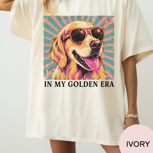 May include: An ivory t-shirt featuring a graphic of a golden retriever wearing red sunglasses. The dog is set against a teal and coral sunburst background. Below the graphic, the text reads IN MY GOLDEN ERA in a bold, black, serif font.