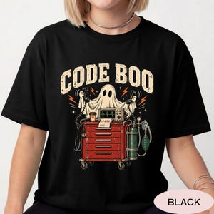 May include: A black t-shirt featuring a graphic of a white ghost holding two telephone handsets above a red medical crash cart. The cart holds a heart monitor, a coffee cup, and an oxygen tank. The text CODE BOO is printed in large beige letters.