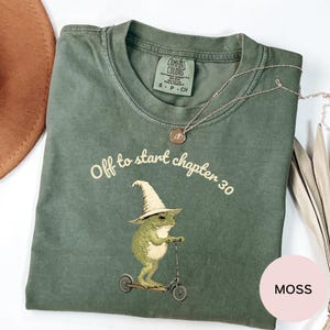 May include: A moss green t-shirt featuring a graphic of a frog wearing a pointed hat and riding a scooter. The text above the frog reads "Off to start chapter 30". The shirt has a crew neckline and a Comfort Colors brand label on the inside collar.