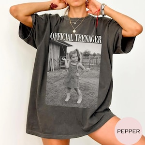 May include: A charcoal gray oversized t-shirt featuring a black and white photographic print of a young child standing in front of a wooden structure. The text "OFFICIAL TEENAGER" is printed in white capital letters above the photograph.