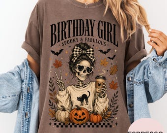 May include: A brown t-shirt featuring a graphic of a skeleton wearing heart-shaped sunglasses and a plaid hair bow. The skeleton holds a coffee cup and makes a peace sign. Text reads BIRTHDAY GIRL SPOOKY & FABULOUS with pumpkins and bats.