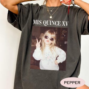 May include: A charcoal gray t-shirt featuring a rectangular graphic print of a young person wearing sunglasses and making a peace sign. The text MIS QUINCE XV is printed in white above the image. The shirt has a crew neckline and short sleeves.