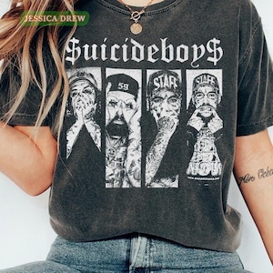 Puede incluir: Camiseta gris oscuro con el nombre de la banda "Suicideboys$" en fuente gótica. La camiseta muestra un gráfico en blanco y negro de cuatro miembros de la banda. La camiseta tiene un aspecto vintage y desgastado.