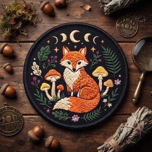May include: A circular embroidered patch featuring a central orange and white fox surrounded by yellow mushrooms, green ferns, and small purple flowers. Above the fox, a crescent moon and stars are stitched in gold thread on a black background.