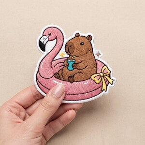 May include: An embroidered patch featuring a brown capybara sitting inside a pink flamingo-shaped pool float. The capybara holds a small blue cup with a yellow straw. A yellow ribbon bow is attached to the side of the float, and a small yellow flower and a star-shaped sparkle are embroidered nearby.