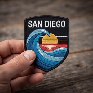 May include: A shield-shaped embroidered patch with a black background and a white border. The design features the text SAN DIEGO in white letters above a large blue ocean wave, a yellow sun, and horizontal lines in red, orange, and yellow.
