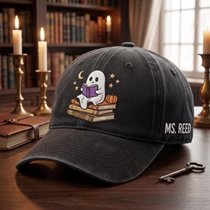 May include: A charcoal gray baseball cap featuring an embroidered graphic of a white ghost sitting on a stack of books and reading a purple book, with a small orange pumpkin, stars, and a crescent moon. The text MS. REED is printed on the side.