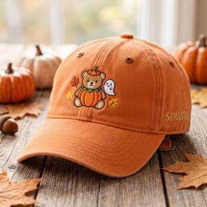 May include: An orange baseball cap featuring an embroidered design of a teddy bear wearing a pumpkin costume, accompanied by a small white ghost and autumn leaves. The name SAMANTHA is embroidered in gold thread on the side of the cap.