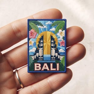 May include: A rectangular embroidered patch featuring a vibrant scene of the Bali gate, a bright yellow sun, blue sky, white clouds, green palm leaves, and colorful hibiscus flowers. The word BALI is stitched in bold, light-colored capital letters at the bottom.