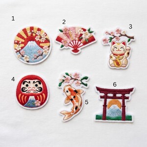 May include: Six embroidered patches featuring Japanese motifs: a circular mountain scene with sun rays, a folding fan with cherry blossoms, a beckoning cat under a branch, a red Daruma doll, a koi fish with blossoms, and a torii gate with a mountain.