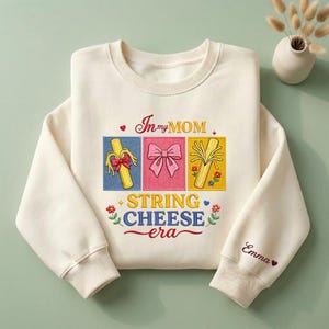 May include: Cream-colored sweatshirt with the text "In my MOM STRING CHEESE era" and three illustrated cheese stick designs. The sleeve cuff has the name "Emma" embroidered. The design includes colorful graphics and floral accents.