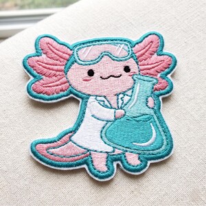 May include: An embroidered patch featuring a light pink axolotl wearing white lab goggles and a white lab coat. The axolotl has dark eyes, a small mouth, and pink feathery gills, and it is holding a large teal-colored glass laboratory flask.