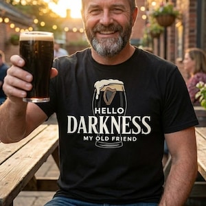 May include: A black t-shirt with the text "Hello Darkness My Old Friend" and a graphic of a pint of stout. The person is holding a pint of dark beer. The scene is outdoors with a wooden table.