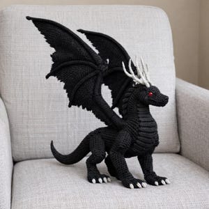 May include: A black crochet dragon with large, spread wings and red eyes. The dragon has white accents on its head and feet, and is posed on a gray armchair. The dragon is a fantasy-themed decorative item.