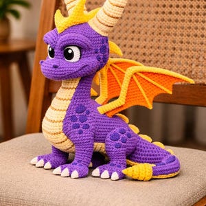 May include: A handmade crochet dragon in shades of purple, yellow, and orange. The dragon has large eyes, horns, and wings. It is sitting on a gray cushion. The dragon is approximately 10 inches tall.