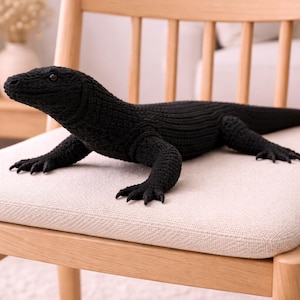Pode incluir: Um brinquedo de lagarto preto tricotado com garras detalhadas e uma cauda longa, repousando sobre uma almofada de cadeira de cor clara. O lagarto é feito de um material texturizado e está posicionado em uma cadeira de madeira.