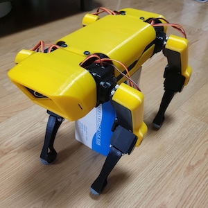 May include: A yellow and black robotic dog with visible wiring and mechanical components. The robot is resting on a wooden floor, with a blue and white box supporting its body. The design suggests a focus on robotics and technology.
