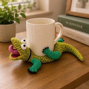 May include: A cream-coloured ceramic mug with a textured herringbone pattern held by a green and yellow crocheted lizard figure. The lizard has a wide-open mouth with a pink tongue, large white eyes, and bright green limbs with black claws.