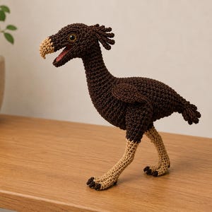 Könnte beinhalten: Eine gehäkelte Vogelfigur mit dunkelbraunem Körper, einem strukturierten Kamm auf dem Kopf und hellbraunen Beinen mit dunkelbraunen Krallen. Der Vogel hat einen gebogenen, hellbraunen Schnabel, ein weit geöffnetes Maul mit rotem Inneren und ein Auge.