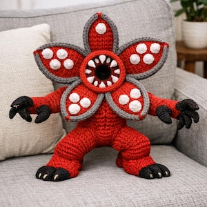 May include: A handmade, red crocheted Demogorgon plush toy with a flower-like head, gray and white accents, and black claws. The creature has a wide-open mouth with white teeth and a black center. It is sitting on a gray couch.