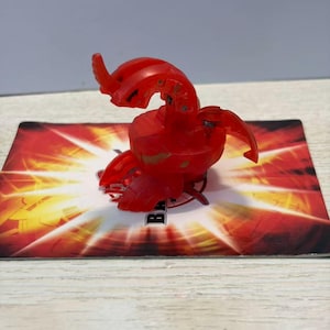 Peut inclure: Une figurine Bakugan en plastique rouge translucide avec une tête incurvée en forme de dragon et des appendices articulés en forme d'ailes. La figurine est placée sur une carte de jeu rectangulaire avec un motif graphique rouge et jaune.