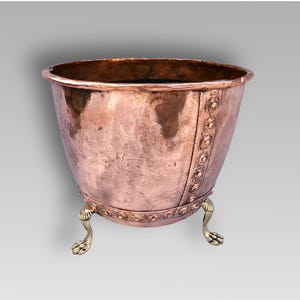 Può includere: Un vaso di rame con un corpo ampio e arrotondato e un bordo leggermente svasato. Il vaso è sostenuto da tre piedini a zampa di leone color ottone. Rivetti decorativi e una fascia in rilievo aggiungono interesse visivo all'esterno.