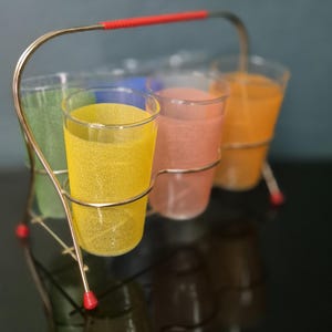 Puede incluir: Un juego de vasos vintage con seis vasos coloridos en un soporte de metal. Los vasos contienen líquidos de varios colores, incluyendo amarillo, rosa y naranja. El soporte tiene un asa y detalles en rojo.