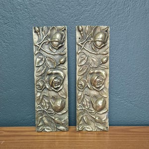 May include: Two rectangular, antique brass panels with a raised floral design featuring roses, leaves, and buds. The panels are vertically oriented and appear to be decorative elements, possibly for furniture or architectural details.