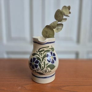 Pode incluir: Um jarro de cerâmica cor de creme com um padrão floral pintado à mão com pétalas azul-escuras e folhas verdes. O jarro tem um pequeno bico e um corpo arredondado, e contém um único ramo de eucalipto seco com folhas redondas.
