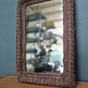 Peut inclure: Miroir rectangulaire en osier marron. Le cadre est en osier tressé et entoure une surface réfléchissante. Le miroir est posé sur une surface en bois contre un mur bleu.