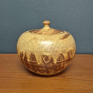 Peut inclure: Un pot en céramique rond et couvert, de couleur jaune clair et marron. Le pot présente une série de motifs triangulaires et géométriques bruns autour de la base. Le couvercle est surmonté d'un petit bouton. Le pot est posé sur une surface en bois.