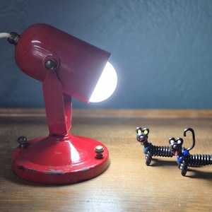 May include: A vintage red desk lamp with a bright light, next to two small, whimsical wire dog sculptures. The lamp has a round base and a pivoting head. The dogs are black with yellow and blue accents.