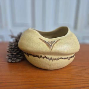 Peut inclure: Un bol en céramique beige avec une forme arrondie et organique et un bord large et ondulé. L'extérieur présente une finition mate et texturée avec une ligne horizontale dentelée brun foncé entourant le milieu et un petit accent en V.